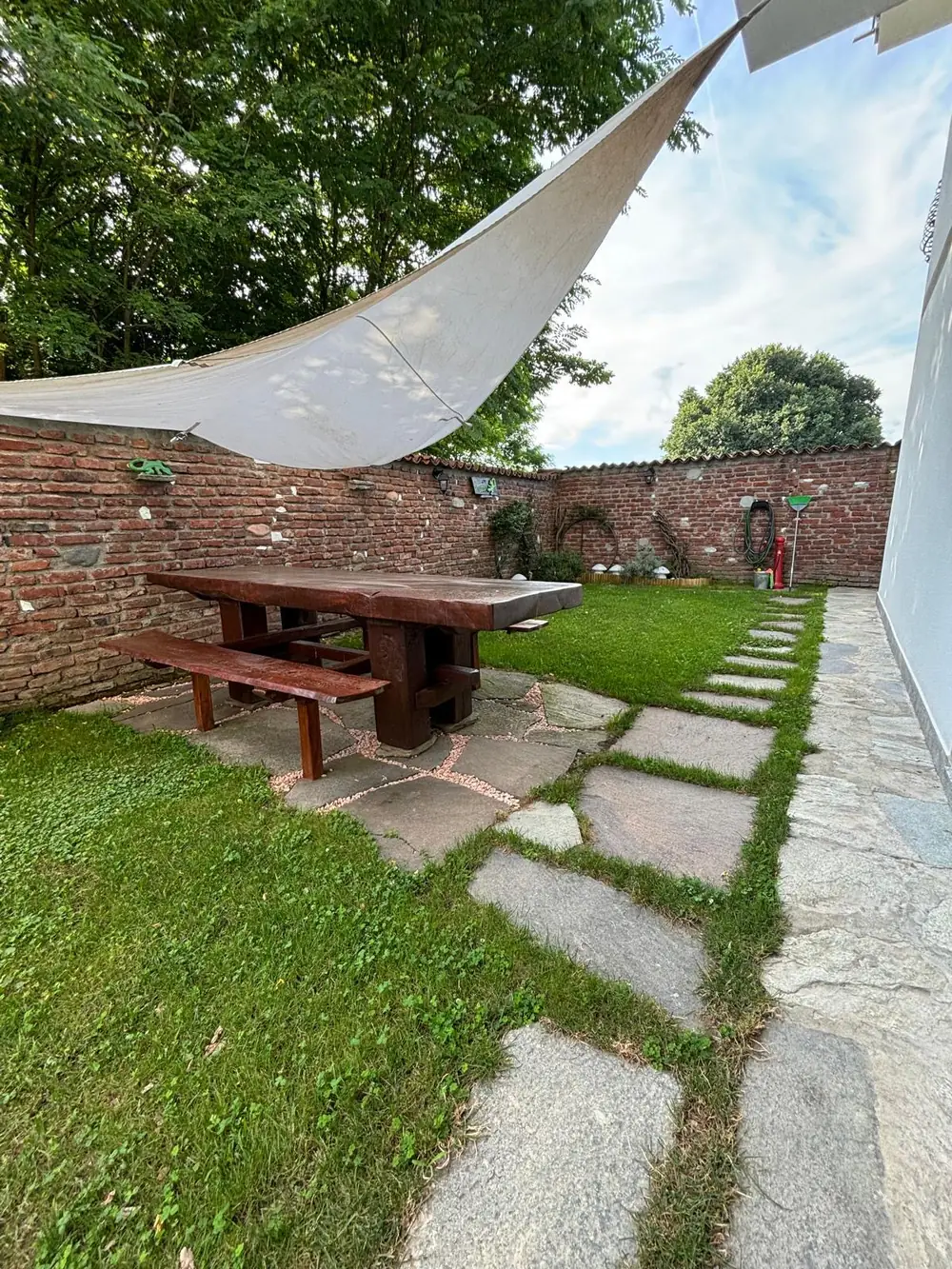 Il giardino con il grande tavolo in legno, la panca e la vela ombreggiante sopra il muro di mattoni